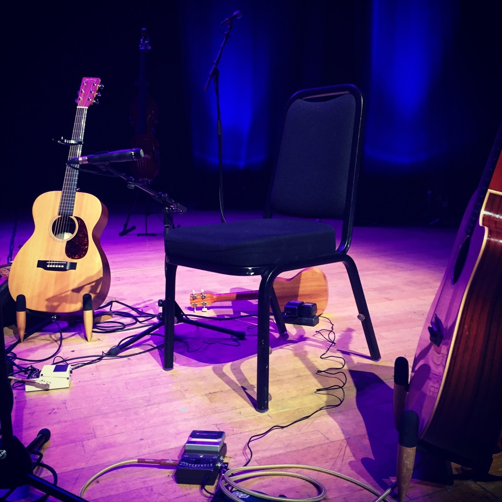 Image of an empty chair, 2 guitars and a ukelele on an empty stage